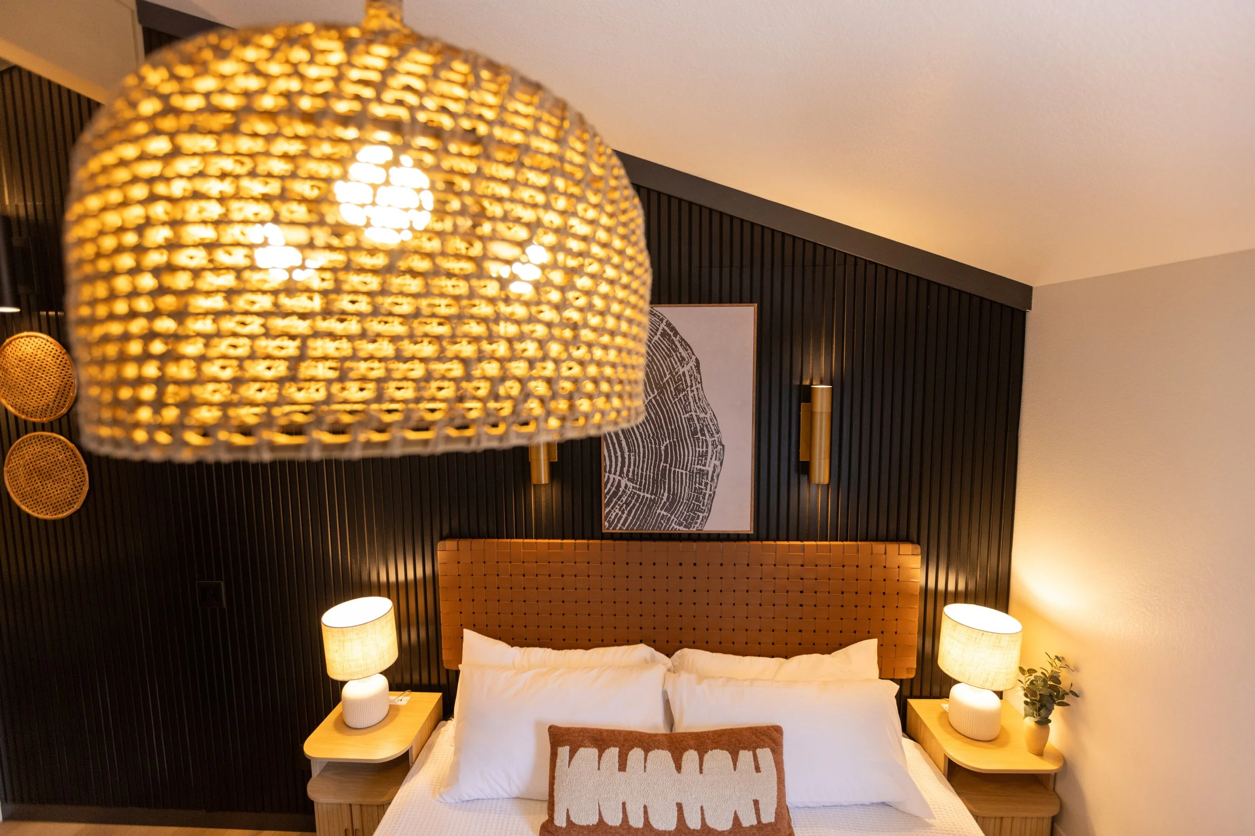 A bedroom with a bed, pillows, and a brown headboard. Nightstands with lamps on each side of the bed. A wall art piece behind the bed. Overhead, a woven hanging light fixture is partially visible.