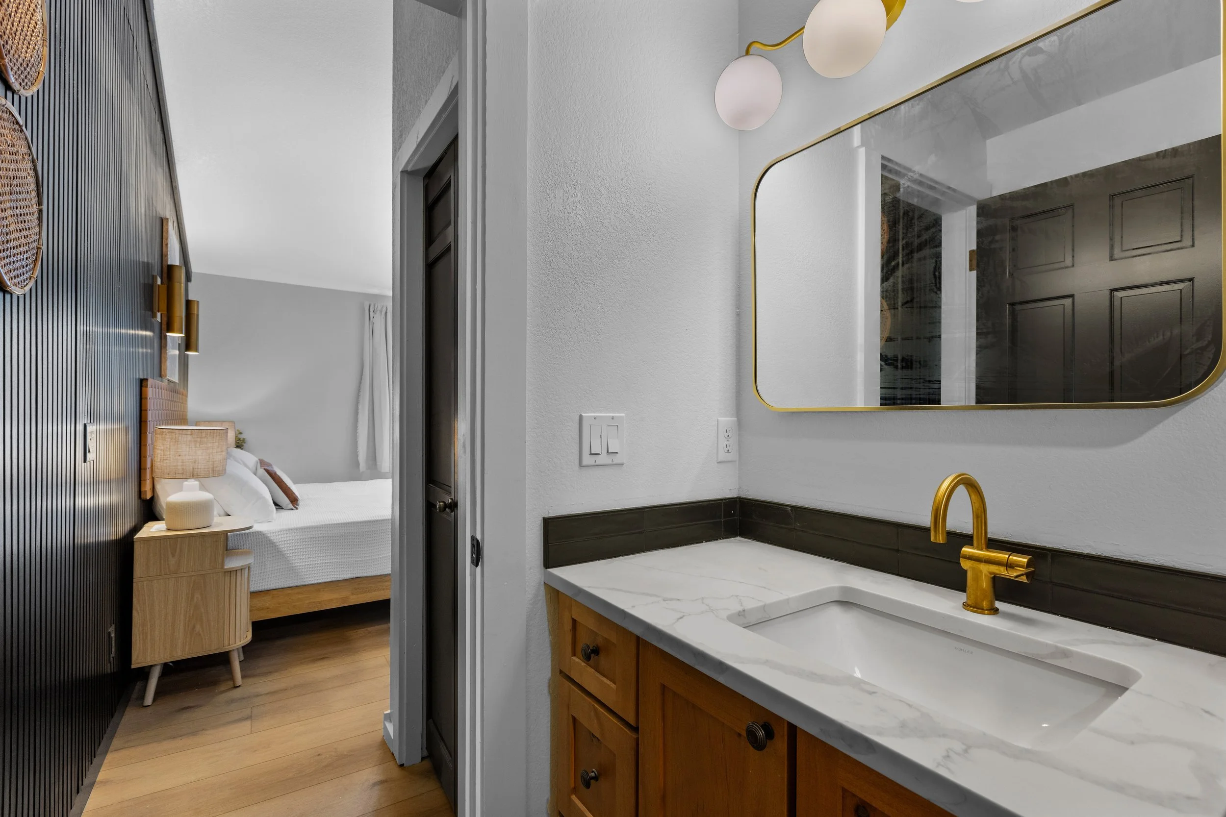 Bathroom vanity with a white marble countertop, a gold faucet, a rectangular mirror with a gold frame, and white spherical light fixtures. Beyond the bathroom, there is a view of a bedroom with a wooden bed frame, white bedding, and a nightstand, wit