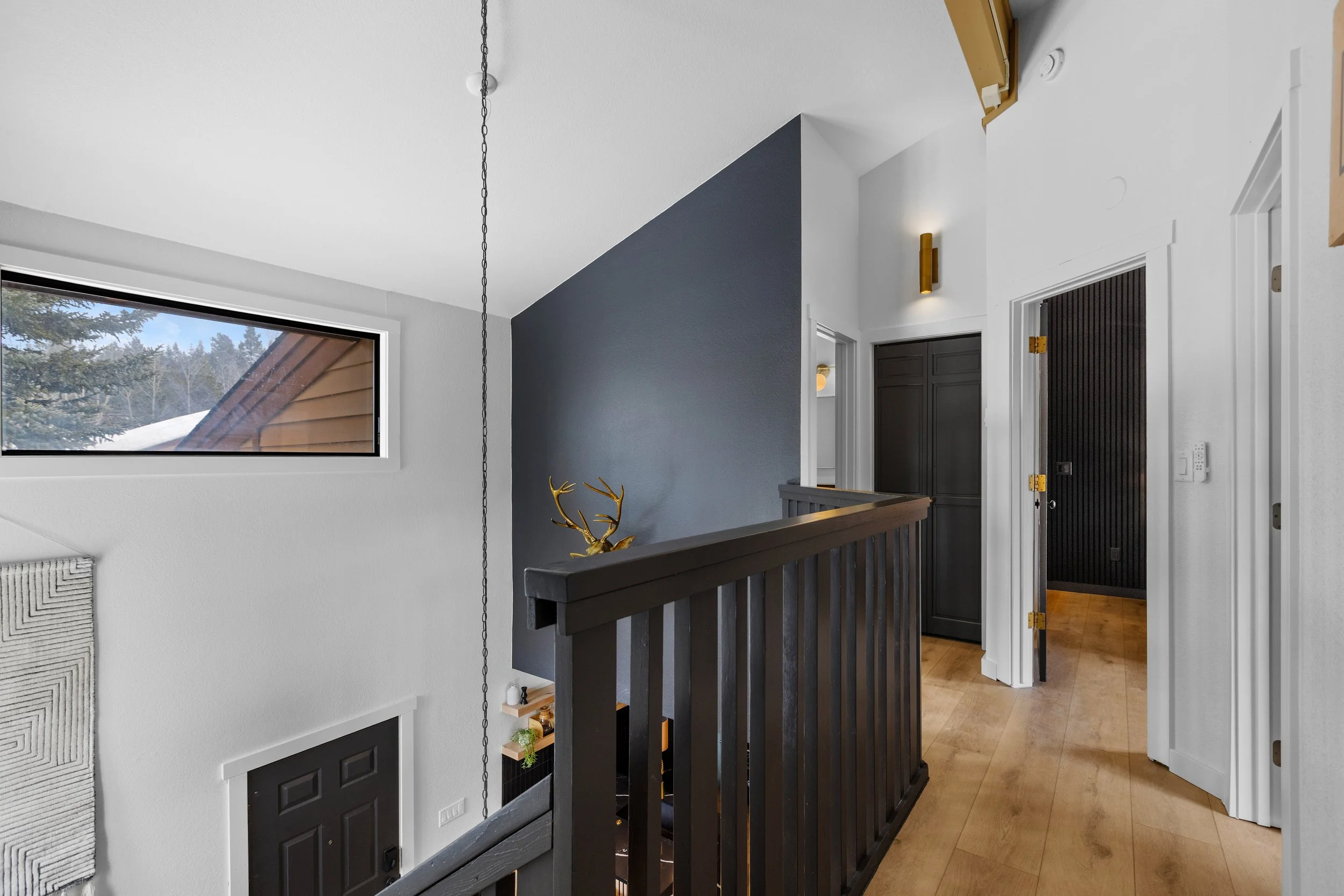 Upper hallway with black wooden railing, dark gray wall, white walls, and a small window showing trees outside.