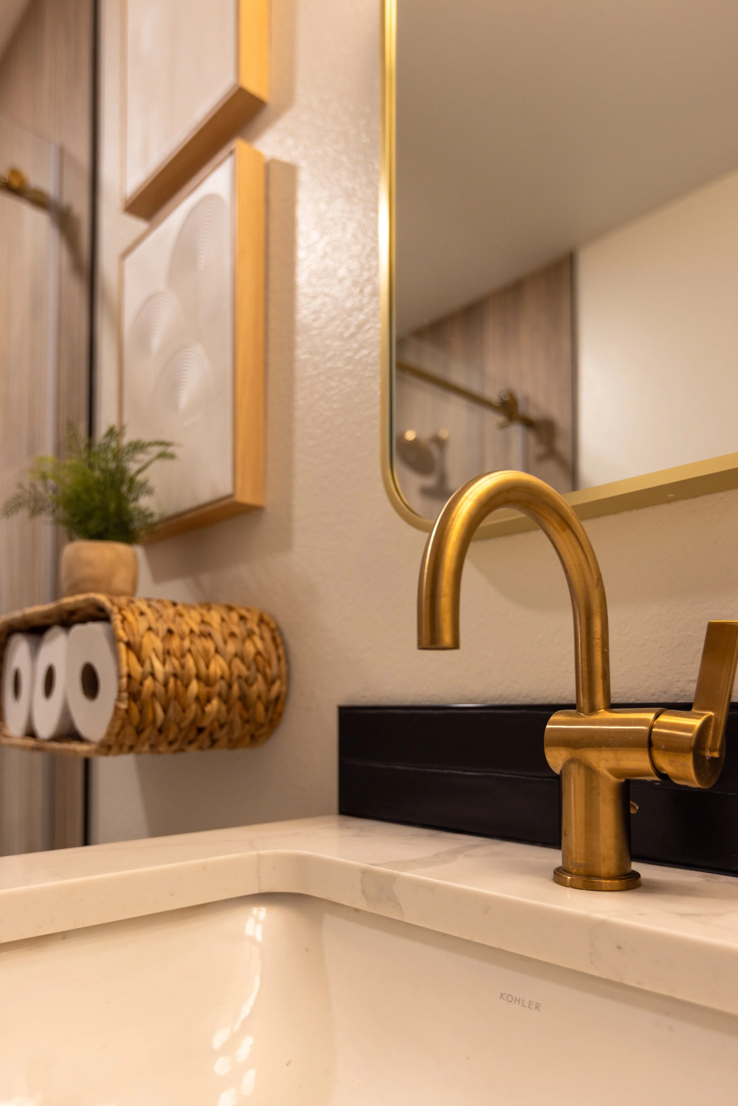 Close-up of a bathroom sink with a gold faucet, a mirror, and a wall-mounted shelf holding toilet paper and a small potted plant.