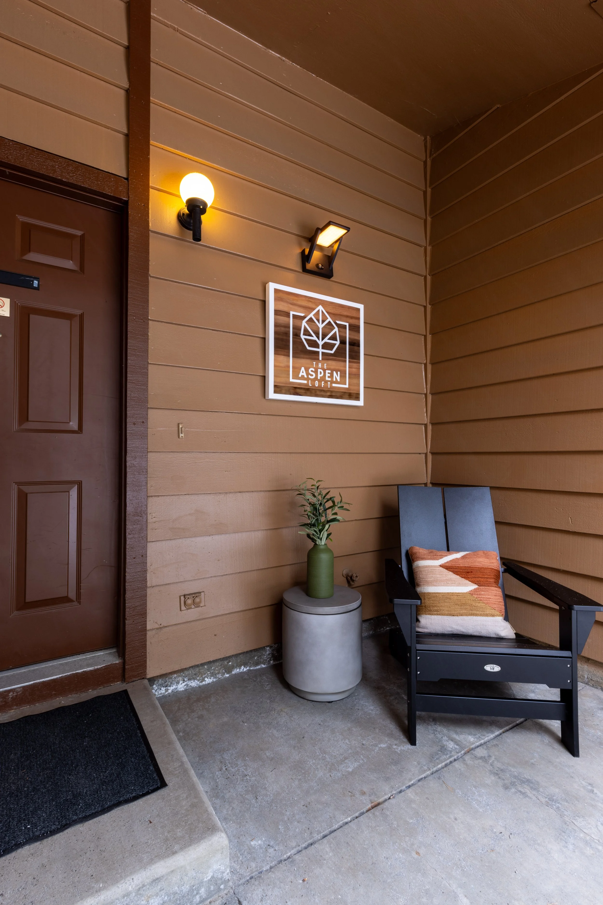 Exterior porch area of The Aspen Loft; brown siding wall with mounted light fixtures, a sign with a leaf logo, a patio chair with a patterned pillow, and a small table with a green vase and plant.