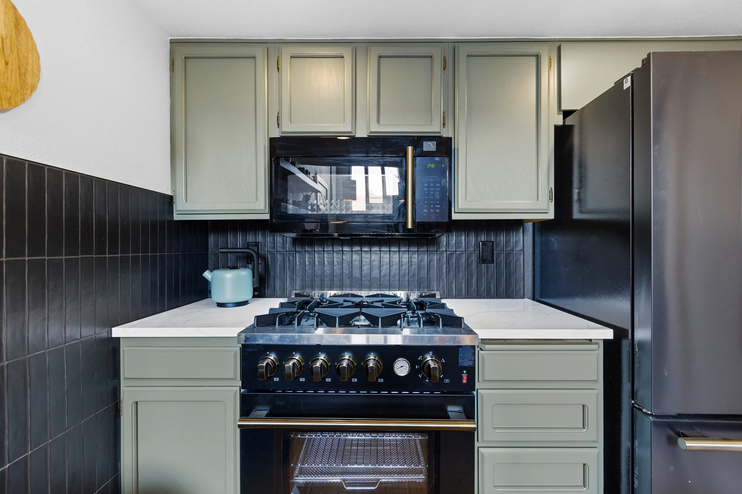 Kitchen with black stove, microwave, green cabinets, black backsplash, and gray refrigerator.