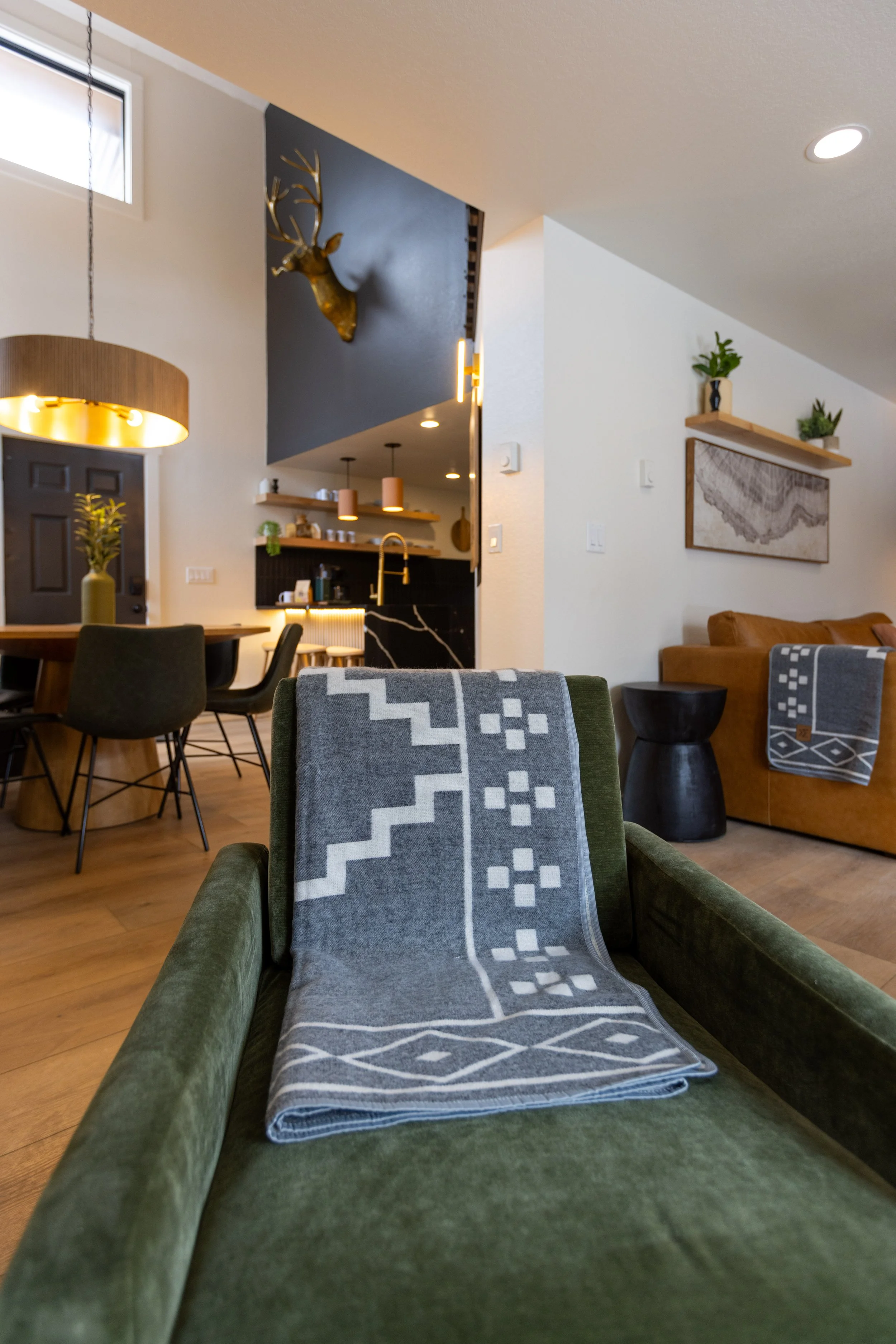 Living room with a green sofa, a gray patterned blanket, a brown leather sofa, wall art, a black side table, and a dining table with black chairs. In the background, a kitchen with black cabinetry and pendant lighting, and a mounted gold-colored deer