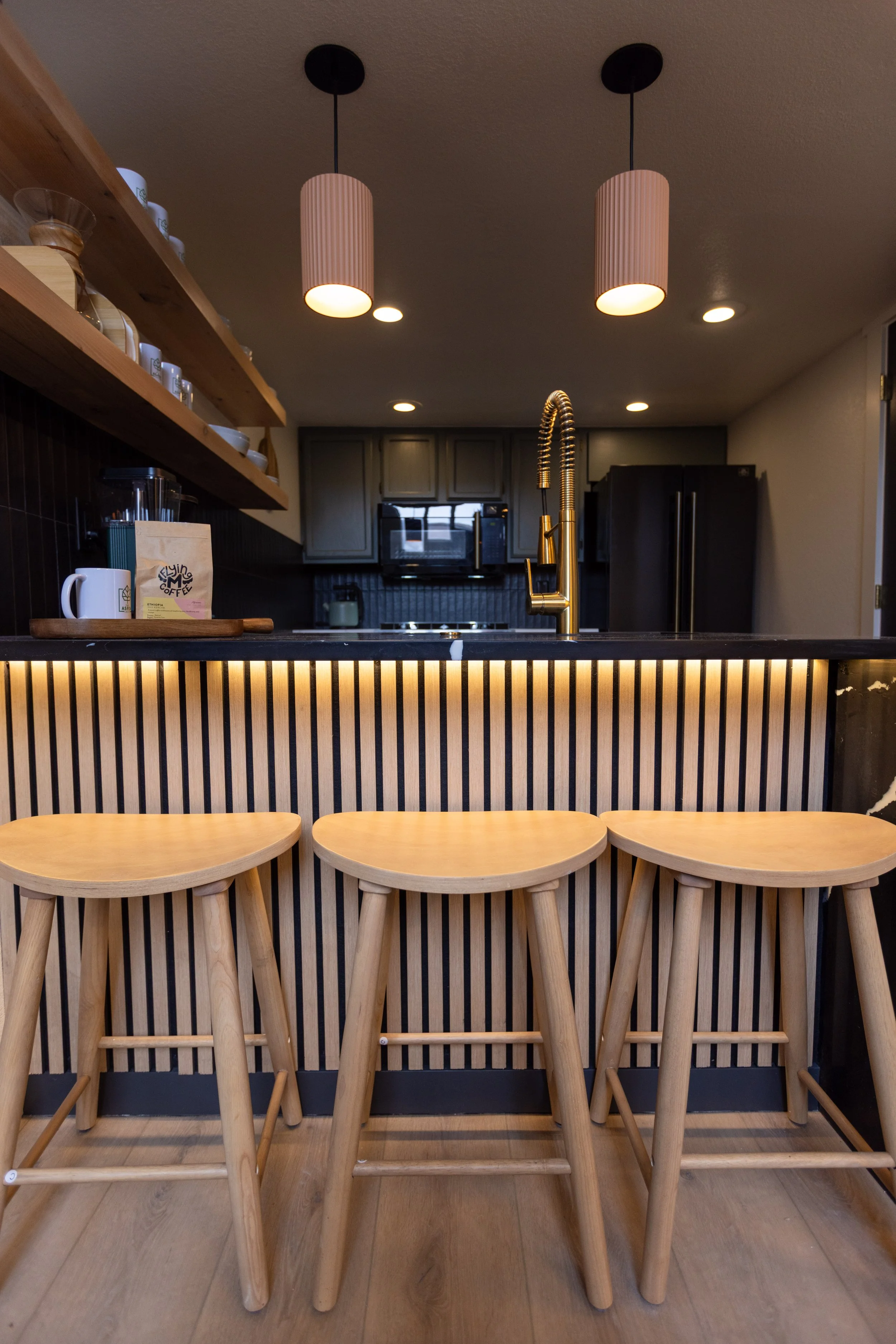 Kitchen with three light wood bar stools in front of a bar counter with vertical black and wood stripes and a gold faucet, black cabinets, and hanging pink pendant lights.