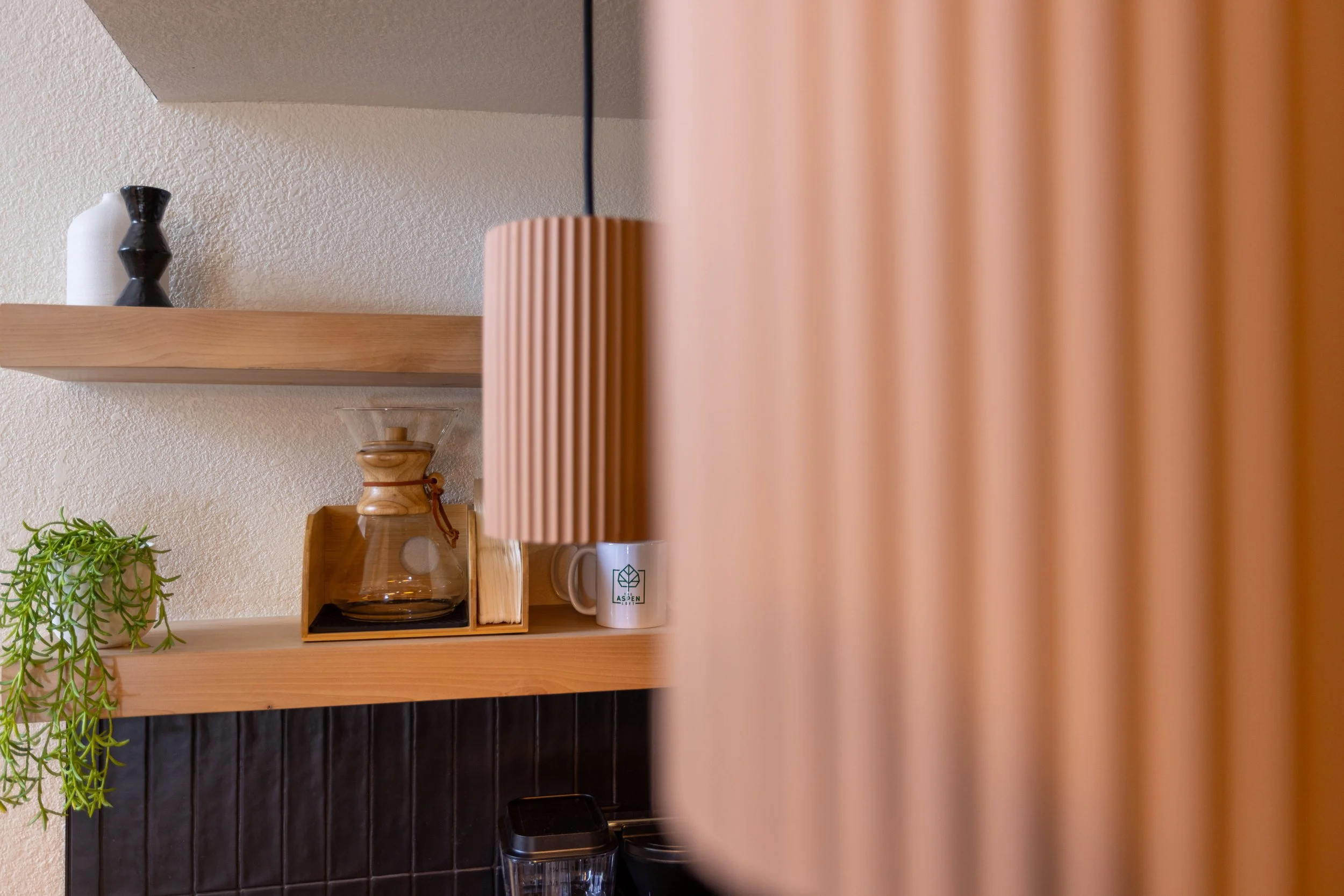 Close-up of a kitchen or counter area with a wooden shelf holding a glass carafe with a wooden lid, a white mug with a leaf logo, a pink ribbed lampshade, and a small potted plant, with a blurred wooden divider in the foreground.
