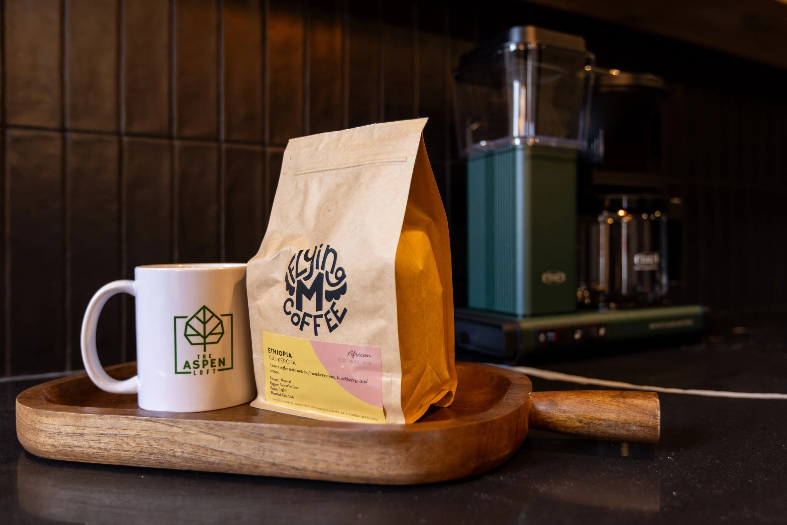A white coffee mug with a green logo reading "The Aspen Loft" next to a brown paper bag of Ethiopia coffee on a wooden tray, with a coffee grinder in the background.