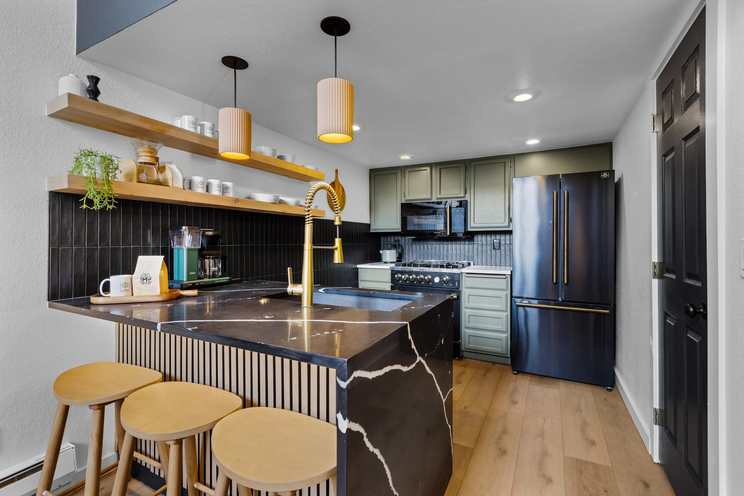 Modern kitchen with black and gold accents, featuring a marble island with a gold faucet, open wooden shelves with dishes and decor, green cabinetry, and black appliances.
