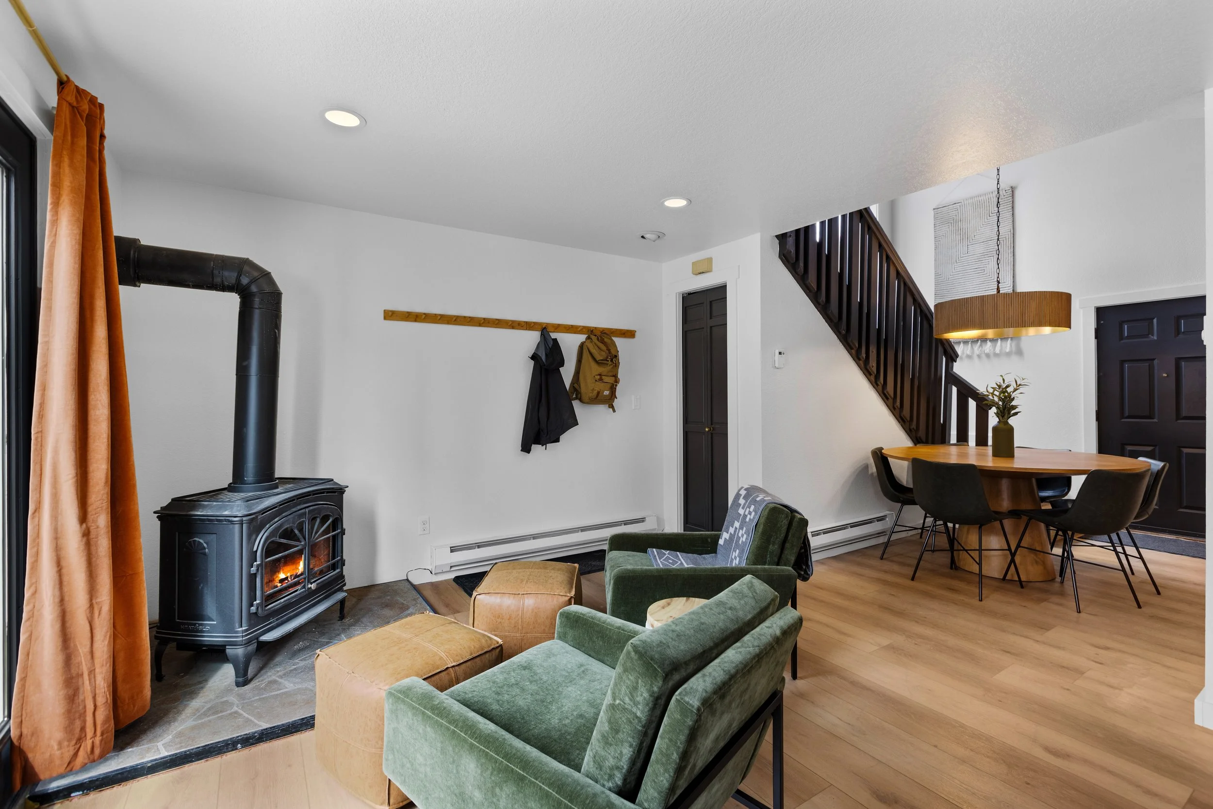 Living room with wood-burning stove, green armchair, small ottomans, round dining table with four black chairs, hanging light fixture, staircase, and natural wood floor.