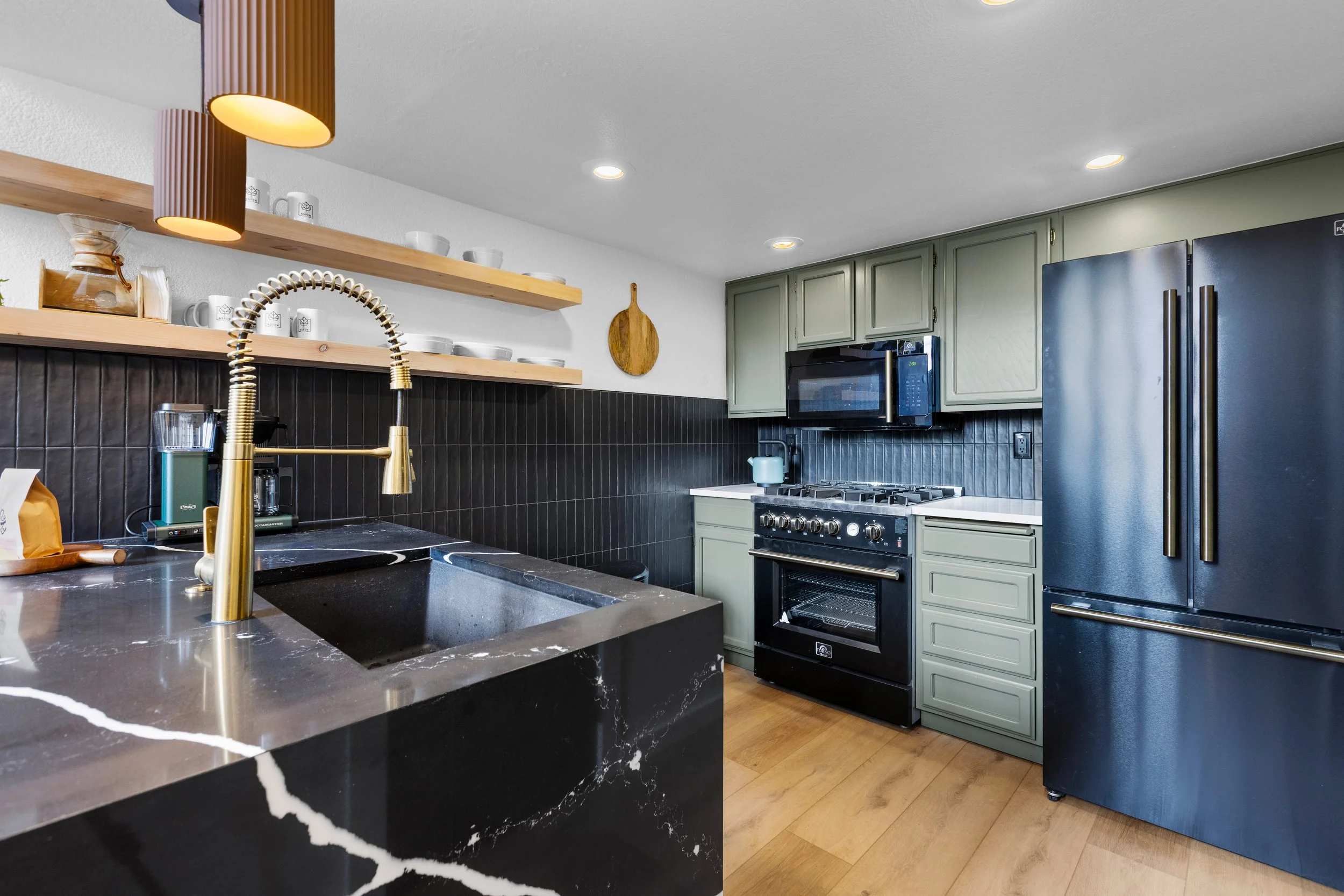 Modern kitchen with black marble countertop and gold faucet, open wooden shelves with white dishes, green cabinets, black appliances, and a black textured backsplash.