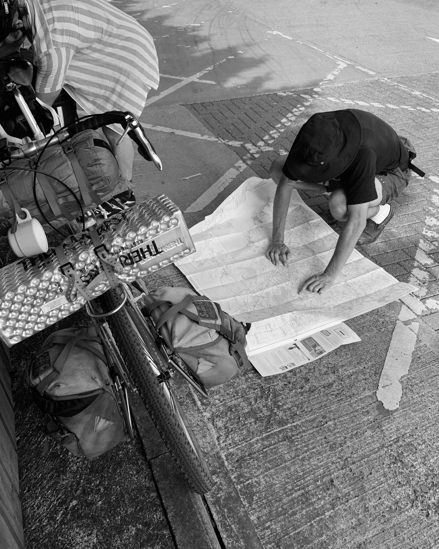 A man studying a large map on the sidewalk, surrounded by bicycles, backpacks, and a plastic bottle, with the scene in black and white.