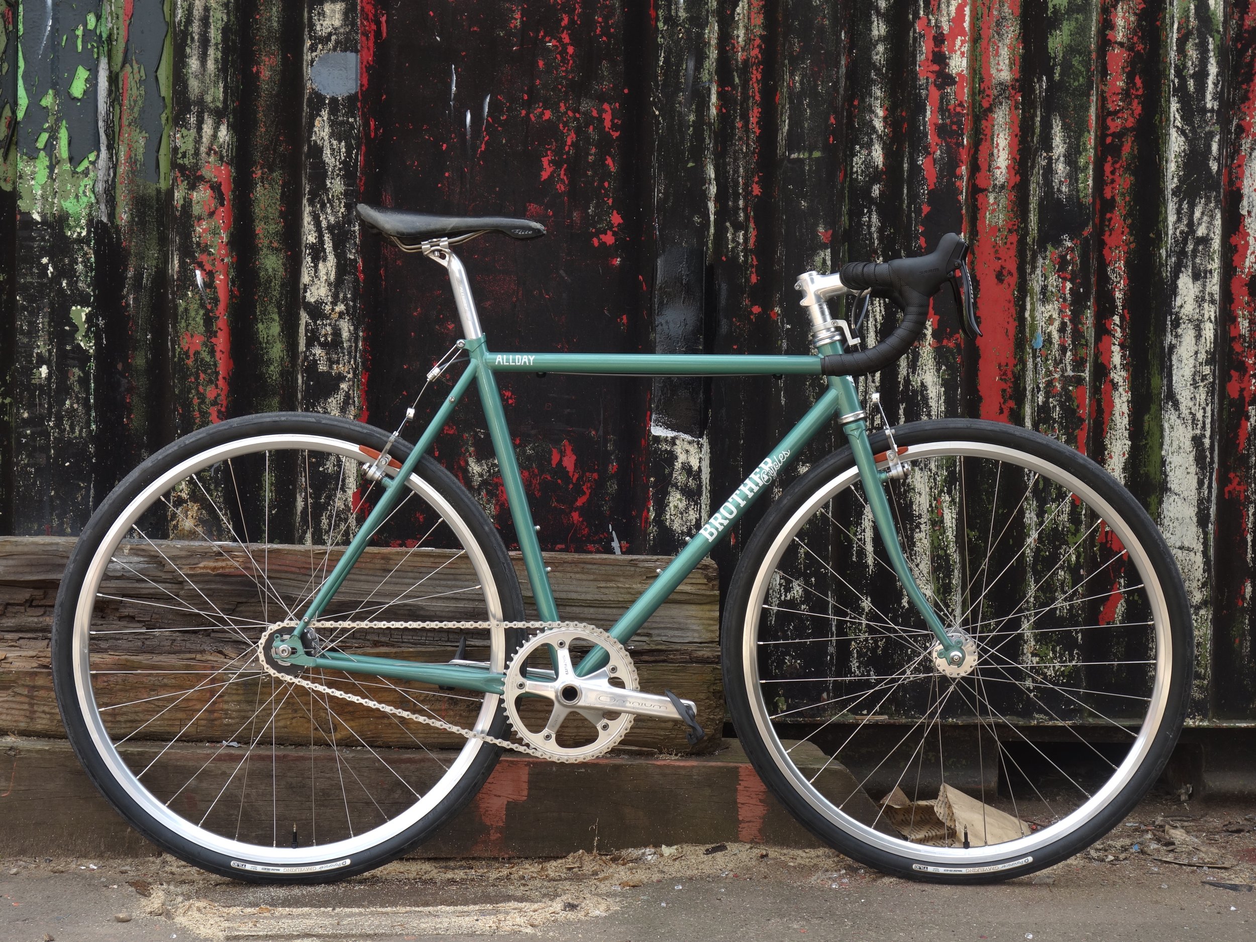 A teal-colored road bicycle with drop handlebars and thin tires, parked in front of a weathered wooden fence with peeling paint.