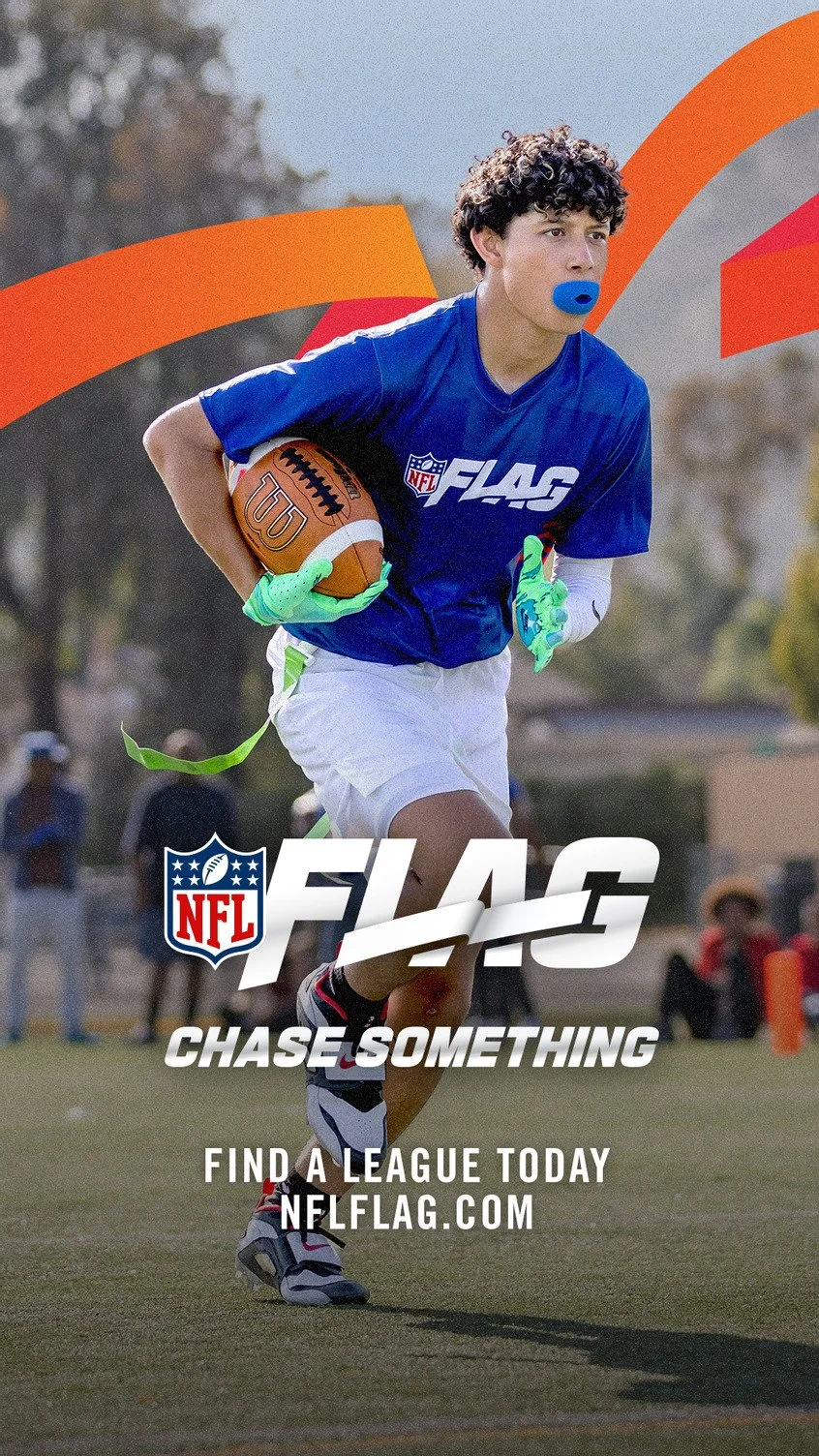 Young male football player running with a football, wearing a blue jersey with NFL FLAG logo, white shorts, green gloves, and a blue mouthguard, on a football field with a blurred background.