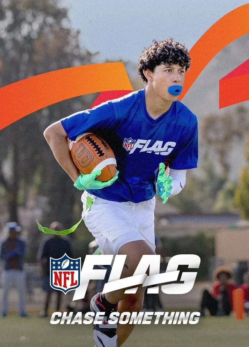 Young male football player running with a football, wearing a blue jersey with NFL FLAG logo, white shorts, green gloves, and a blue mouthguard, on a football field with a blurred background.