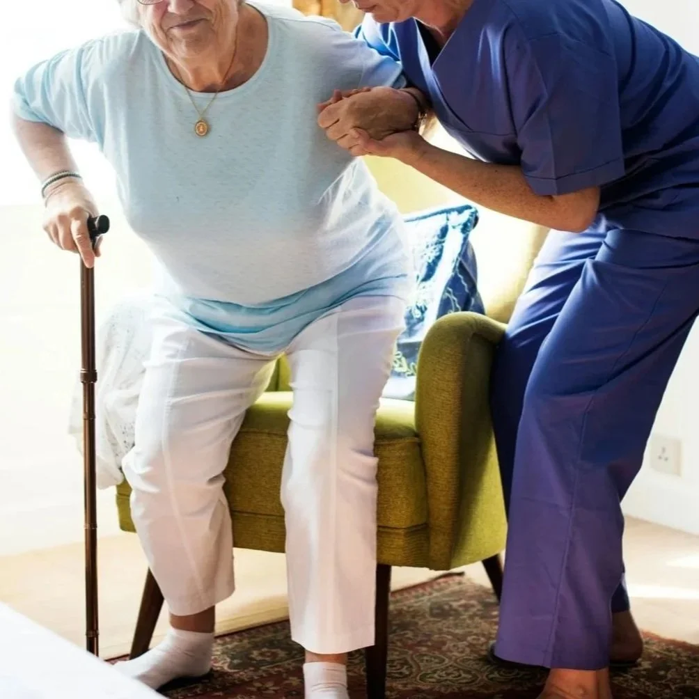 A caregiver assists an older adult from a chair as part of senior respite services at home.