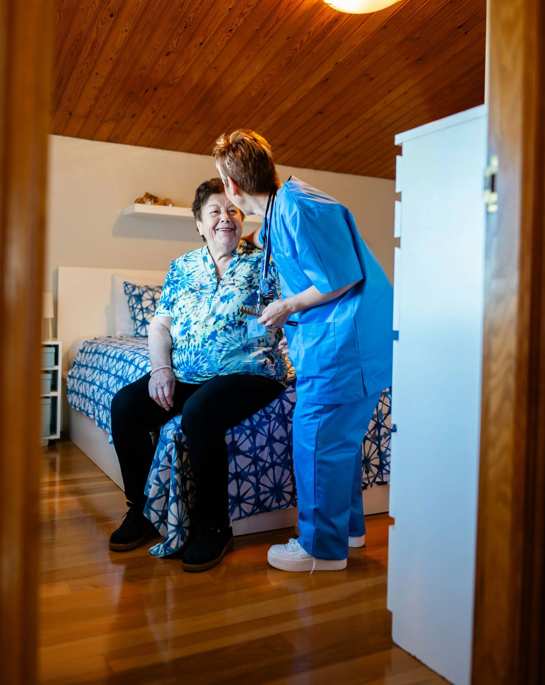 Professional caregiver in blue scrubs assisting an elderly woman in her home.