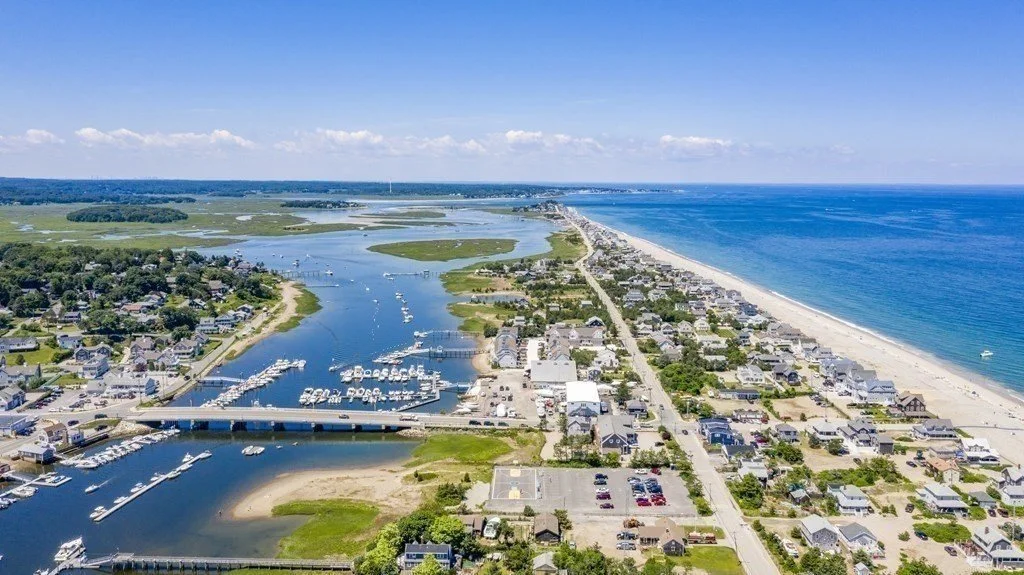 Aerial view of a coastal town with a marina, houses, and a beach along the ocean under a clear blue sky.