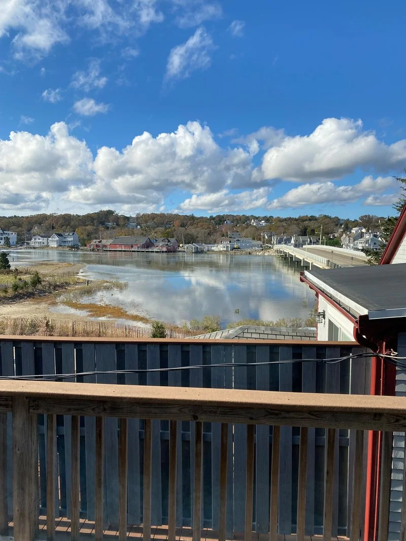 View of a small town over a river with a bridge, houses, and trees, under a partly cloudy sky, taken from a wooden balcony.