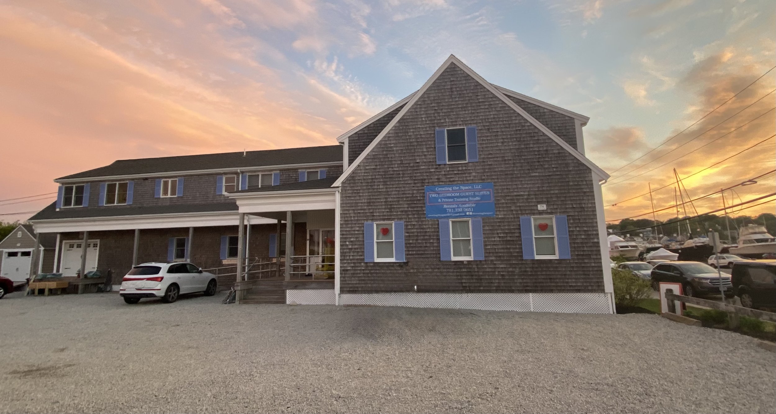 A multi-story building with gray wooden shingles, blue shutters, and a sign advertising two-bedroom guest suites at sunset. There are parked cars in front and boats in the marina to the right. The sky is colorful with pink, orange, and blue clouds.