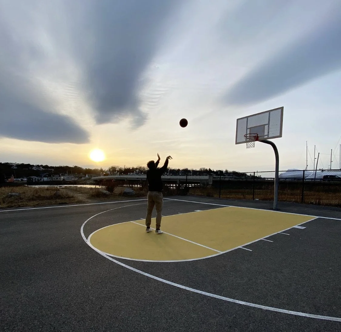 A person shooting a basketball on an outdoor court at sunset, with a single basketball mid-air near the hoop.