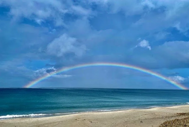 A rainbow over the ocean with a cloudy sky and sandy beach in the foreground.