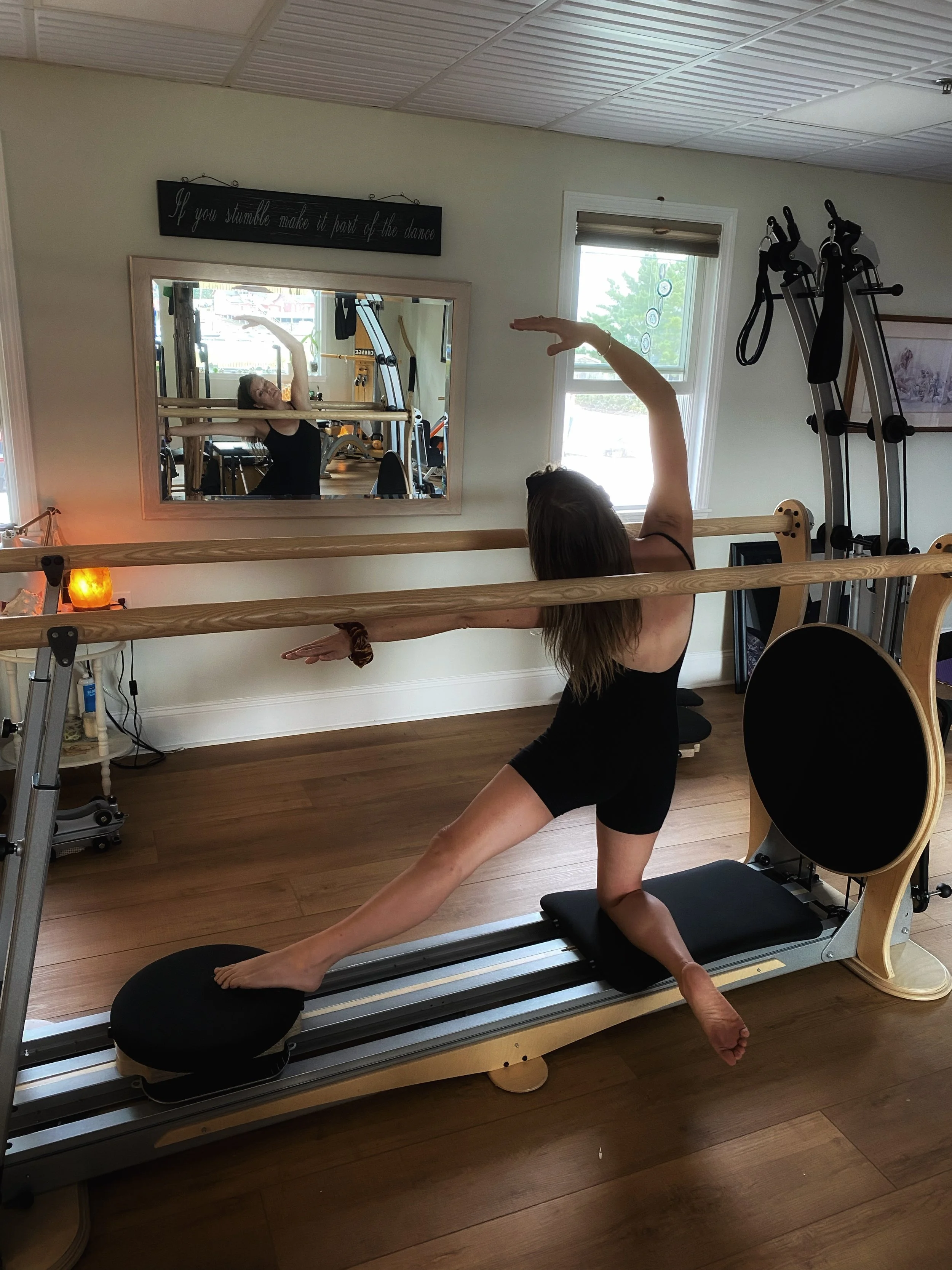 A woman performing a yoga pose on a Pilates reformer machine in a fitness studio, with another woman stretching in front of a mirror.