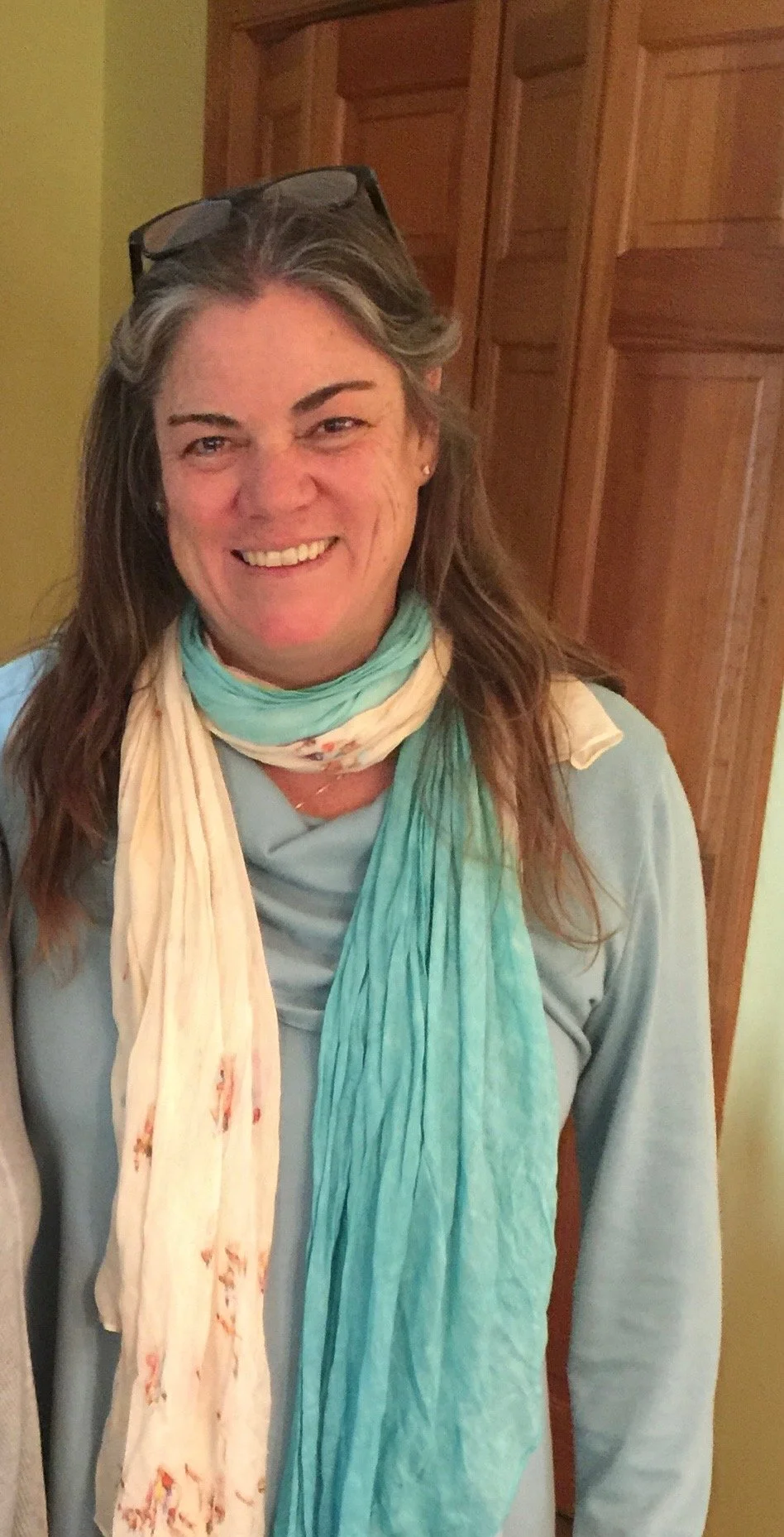 Close-up of a smiling woman with glasses on her head, wearing a light blue top and a scarf around her neck, standing indoors with wooden cabinets in the background.