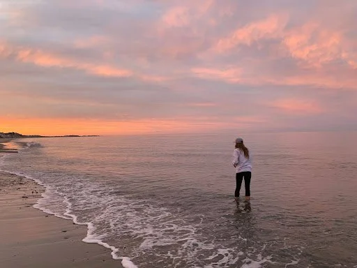 A person standing in shallow water at the beach during sunset, looking out at the horizon with pink and orange clouds in the sky.