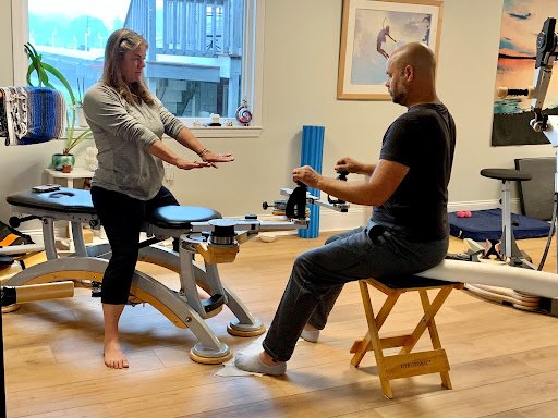 A woman and a man are in a physical therapy or rehabilitation setting. The woman is stretching her leg on a padded table, while the man, sitting on a bench, is assisting or guiding her with therapy exercises. The room has an exercise machine, framed 