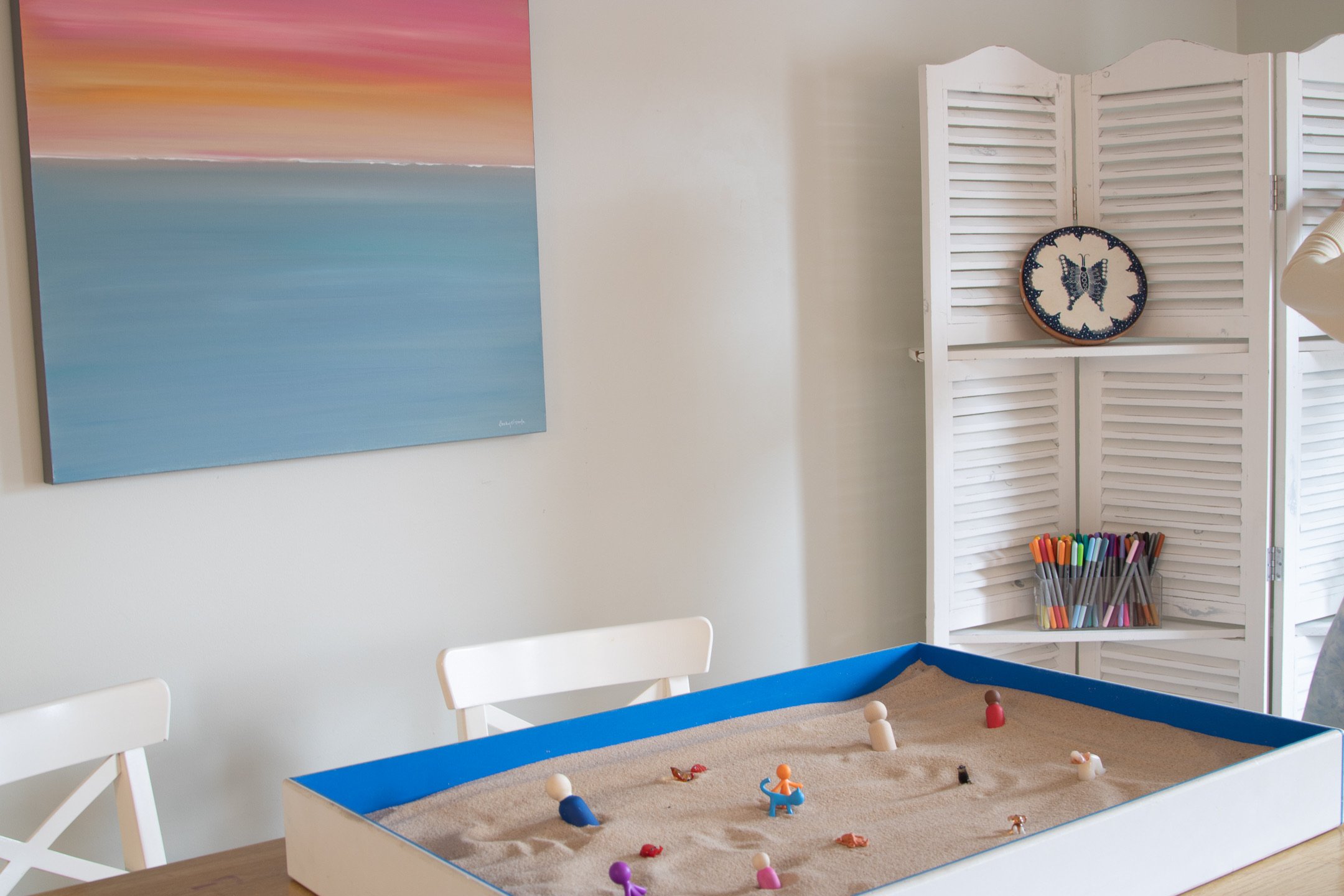 Child's indoor play area with a sandbox filled with sand, small toys, and figurines, surrounded by white chairs and a blue border, in front of a white louvered room divider with a decorative plate featuring a butterfly design, and a large abstract sunset painting on the wall.