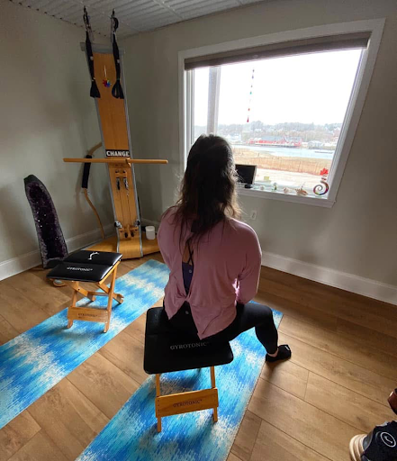 Woman seated on a stool, facing a large window in a room with light-colored walls and wooden flooring, with fitness equipment and a chair nearby.