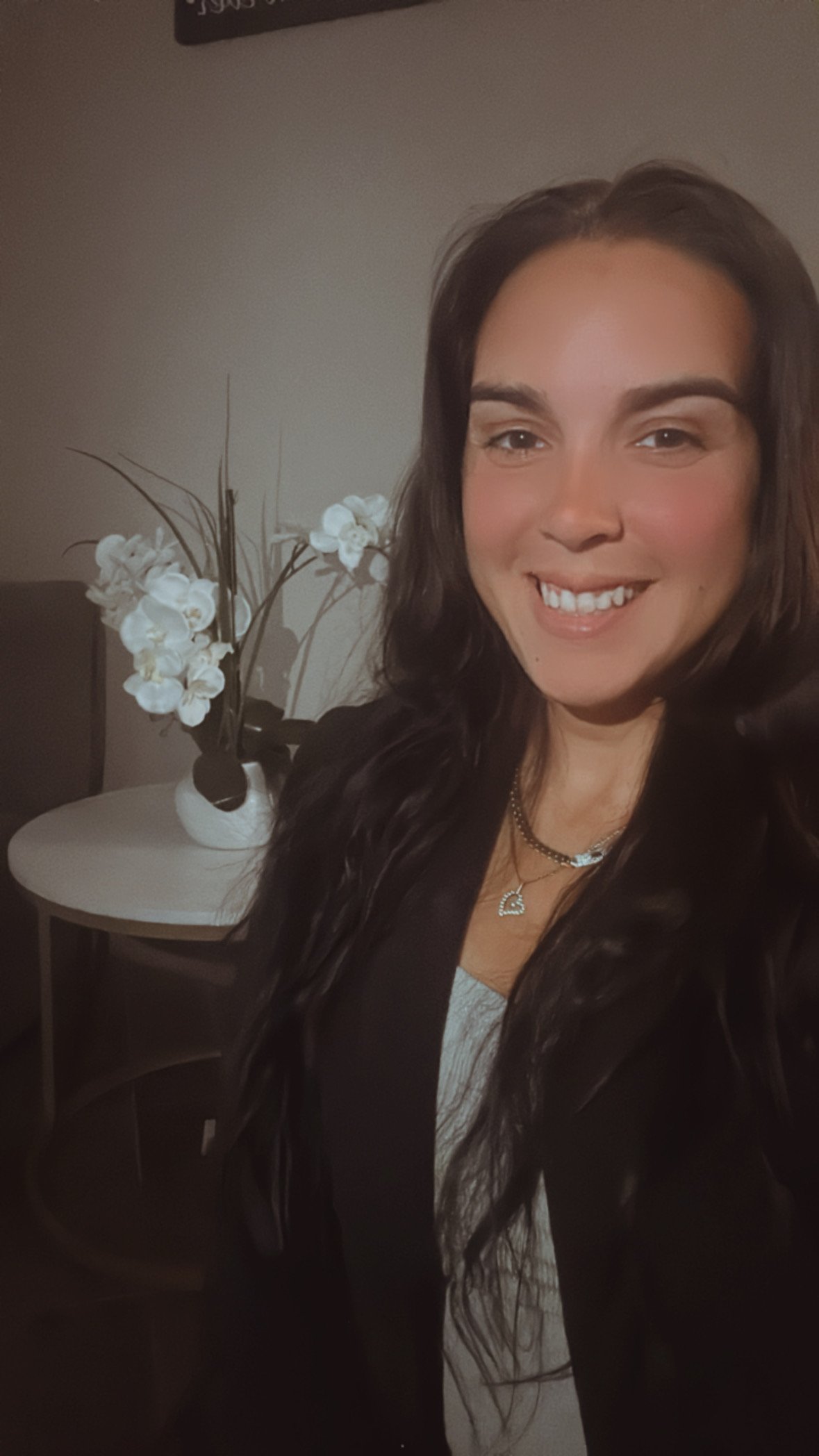 A woman taking a selfie indoors, standing next to a small white table with a white flower vase holding white orchids and decorative branches.