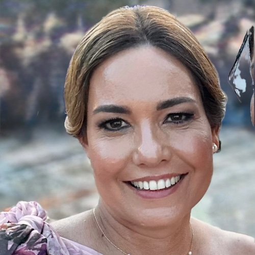 A smiling woman with short brown hair, wearing pearl earrings, outdoors near water with blurred rocks in the background.
