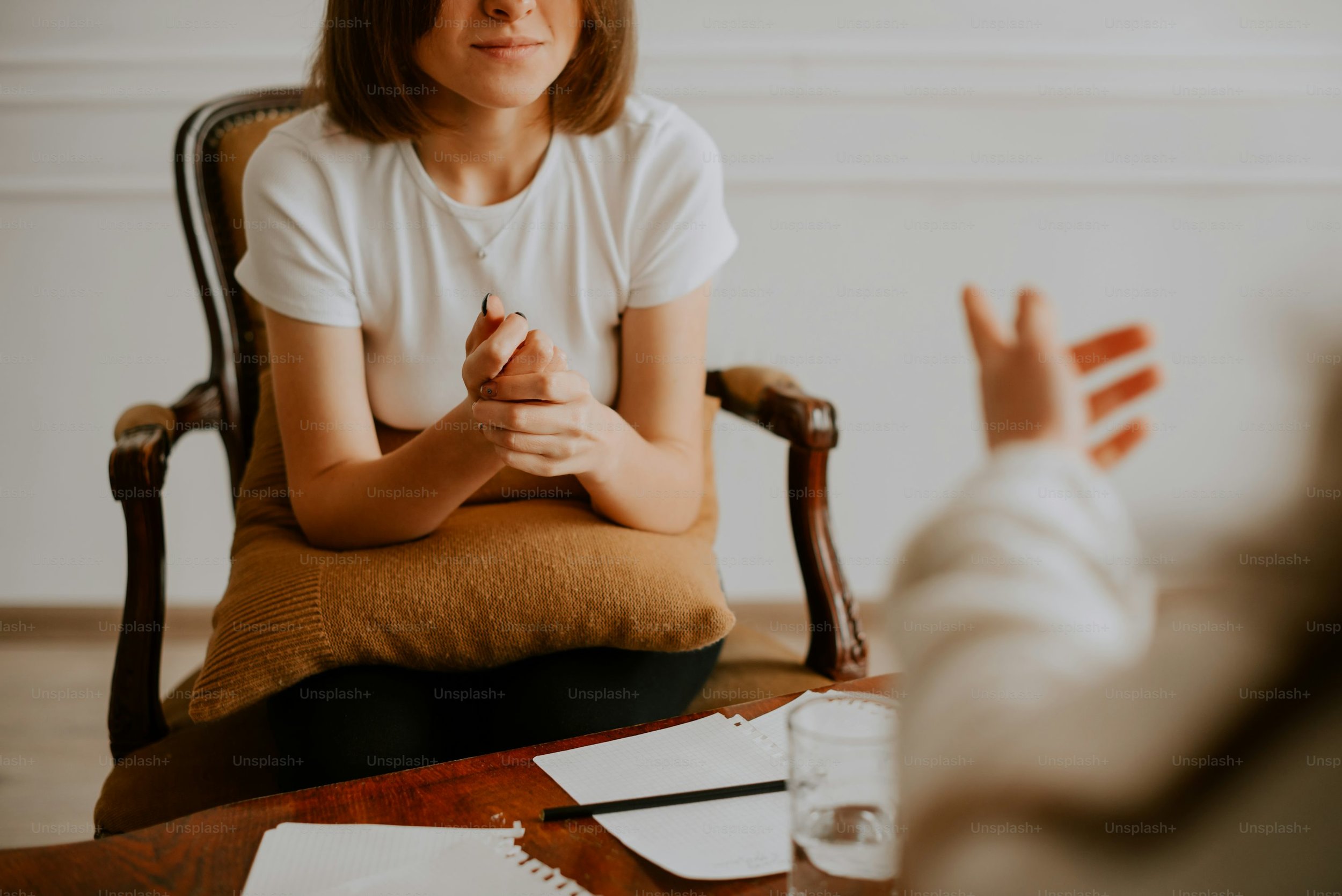A woman sitting on a chair with a cushion on her lap, engaging in a conversation with a person whose hand is gesturing. The scene appears to be in a casual setting, possibly during a discussion or counseling session.