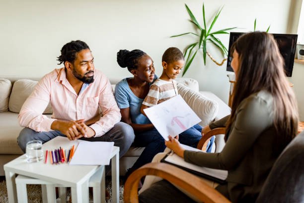 A family of four sitting on a couch during a therapy session with a counselor in a living room, with a potted plant in the background.