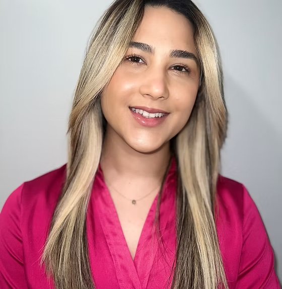 A young woman with long blond hair, wearing a bright pink top, smiling at the camera against a plain light background.