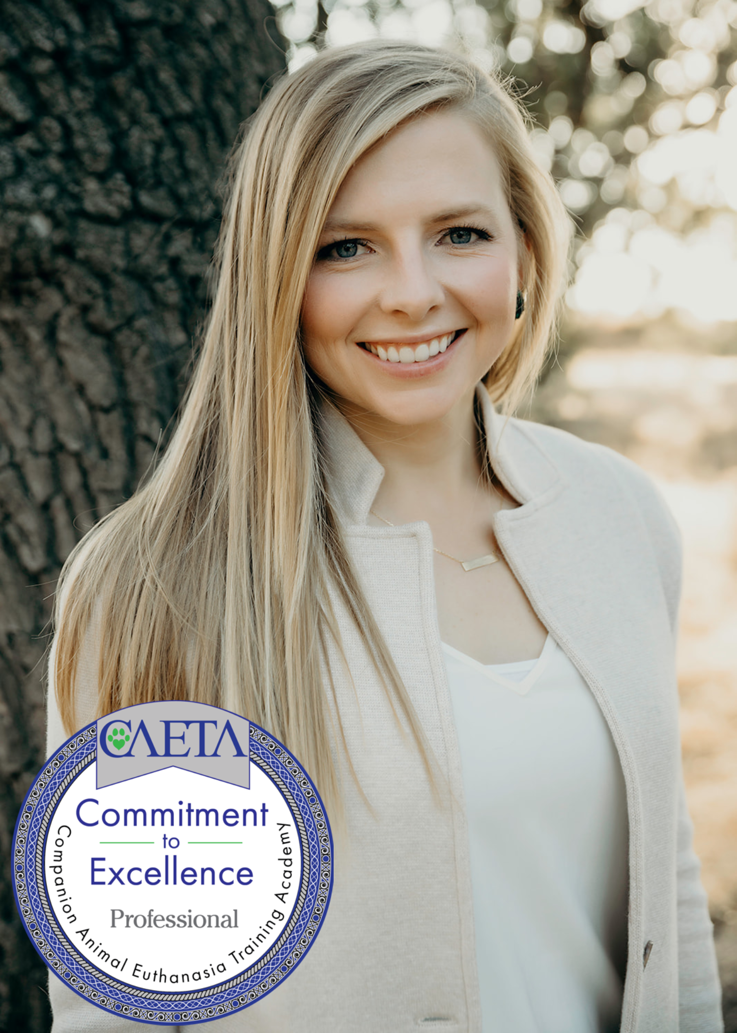 A smiling woman outdoors with sunlight filtering through trees in the background. She has long blonde hair, blue eyes, and is wearing a light-colored blazer. A circular seal is partially visible in the lower left corner, indicating an association with a professional or academic organization related to animal euthanasia training.