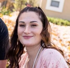 A young woman with long brown hair smiling outdoors, wearing a pink top and a pearl necklace.