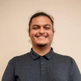 A young man with long hair tied back, smiling, wearing a dark polo shirt, standing against a plain beige background.