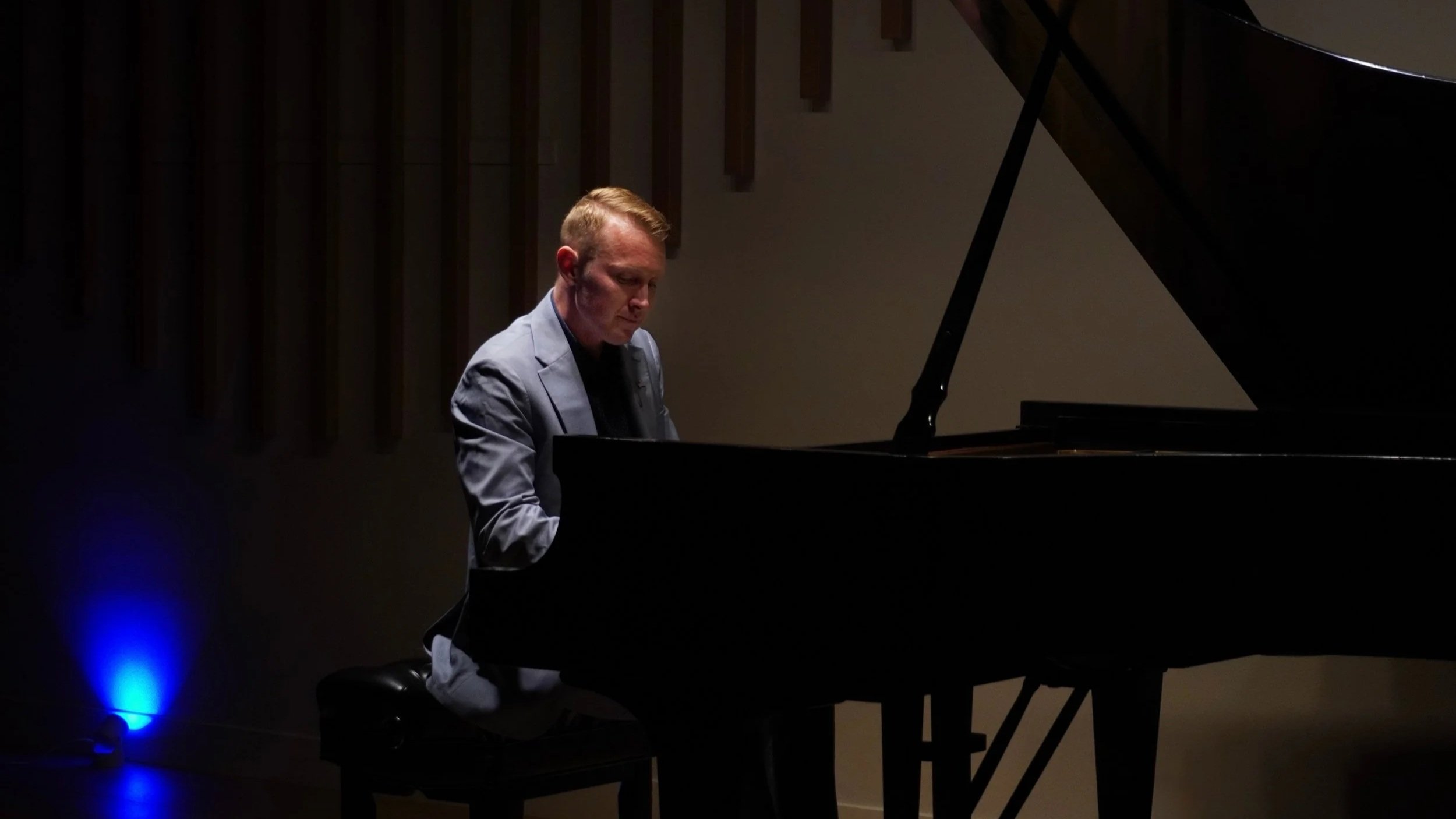 A man in a gray suit playing a grand piano in a dimly lit room.