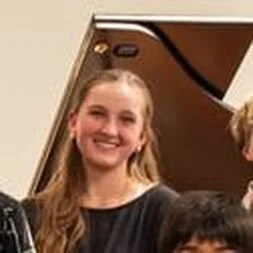 A smiling young girl with long blonde hair standing near a piano.