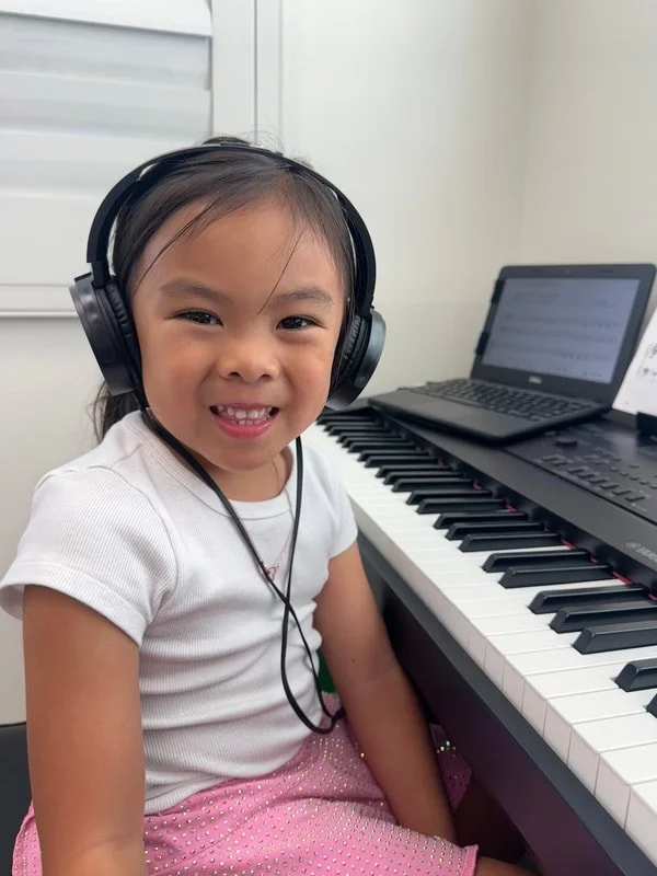 A young girl wearing headphones sitting at a piano, smiling at the camera, with a laptop displaying sheet music nearby.