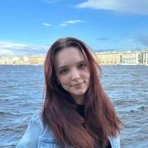 Young woman with long reddish-brown hair smiling by a river with historic buildings and a blue sky in the background.