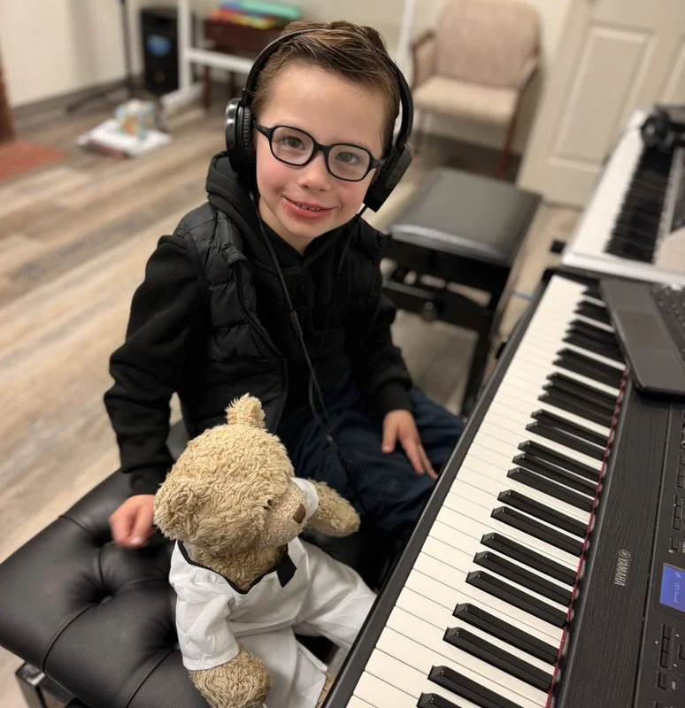 A young boy wearing glasses, a black hoodie, and headphones sitting at a digital piano with a teddy bear dressed as a martial artist in a white gi. The room has wooden floors and some furniture in the background.