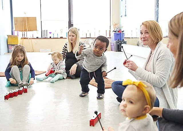 Children and adults in a music classroom sitting and playing with rhythm sticks.