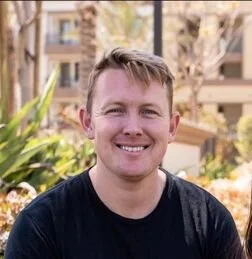 A smiling man with short blonde hair wearing a black t-shirt outside with buildings and trees in the background.