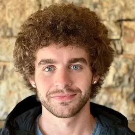 A young man with curly brown hair, blue eyes, and a slight smile, wearing a dark jacket in front of a stone wall.