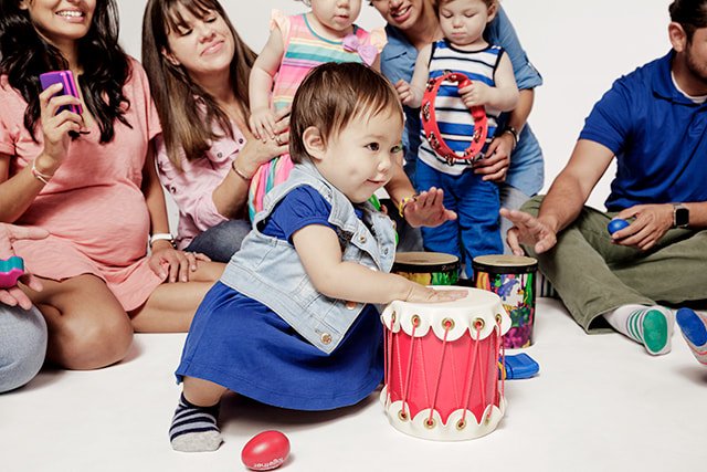A group of children and adults sitting on the floor, with a young boy in a blue dress and denim vest playing a red drum in the center.