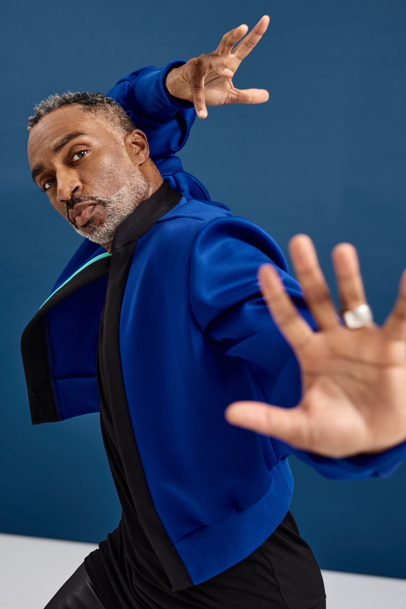 Man with salt-and-pepper hair and beard in a motion pose, wearing a blue jacket, against a blue backdrop.