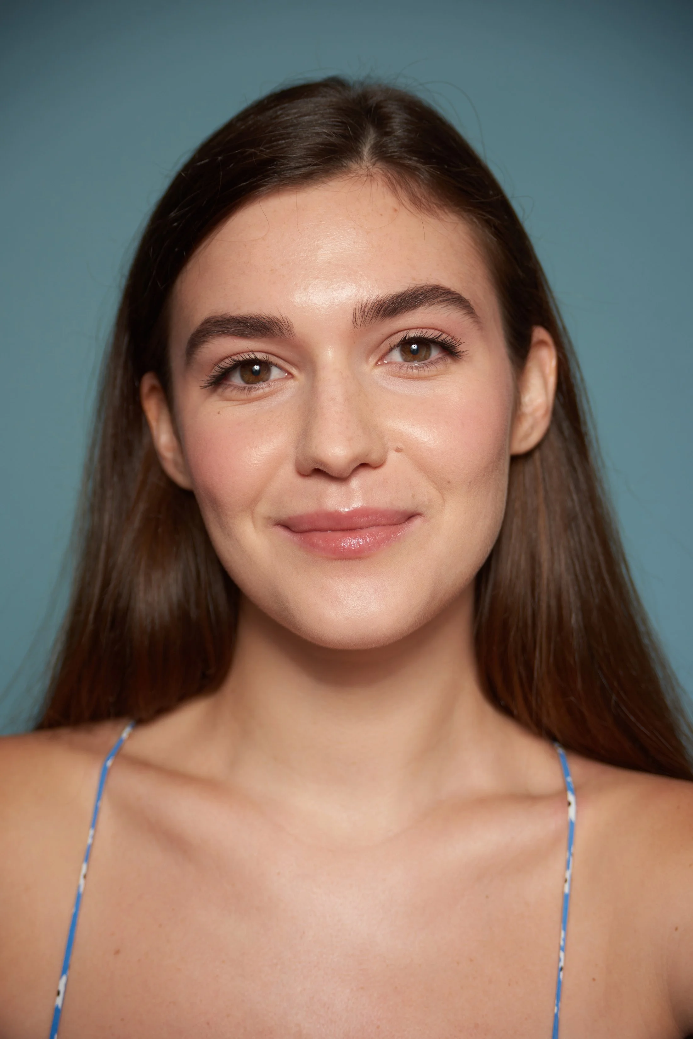 Close-up of a young woman with fair skin, brown hair, and brown eyes, smiling softly against a blue background, wearing a spaghetti strap top.