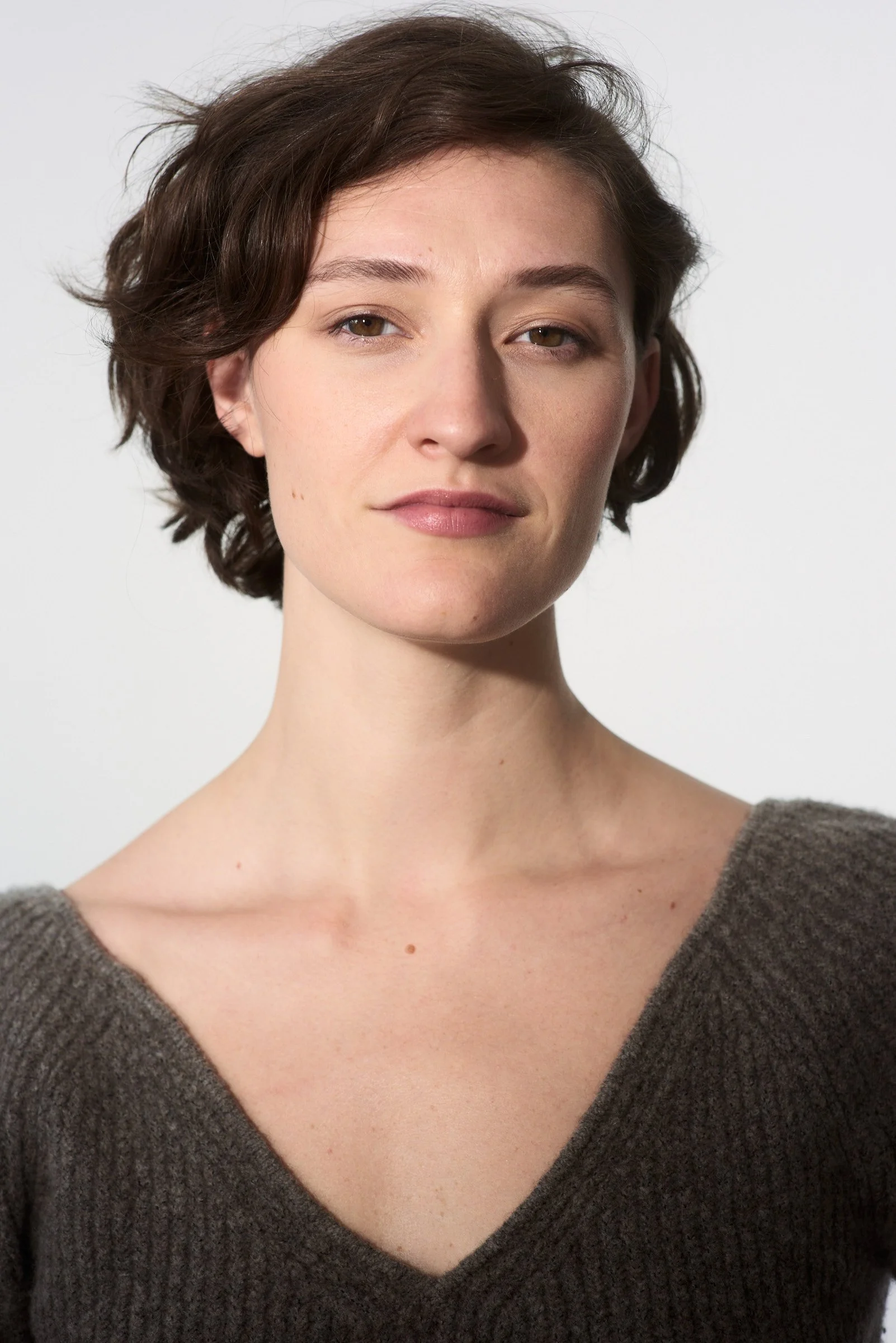 Close-up portrait of a young woman with short brown hair, wearing a gray V-neck sweater, against a plain white background.